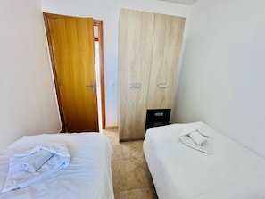 Quarto com duas camas de solteiro, um guarda-roupa de madeira e uma porta que dá para outro espaço.