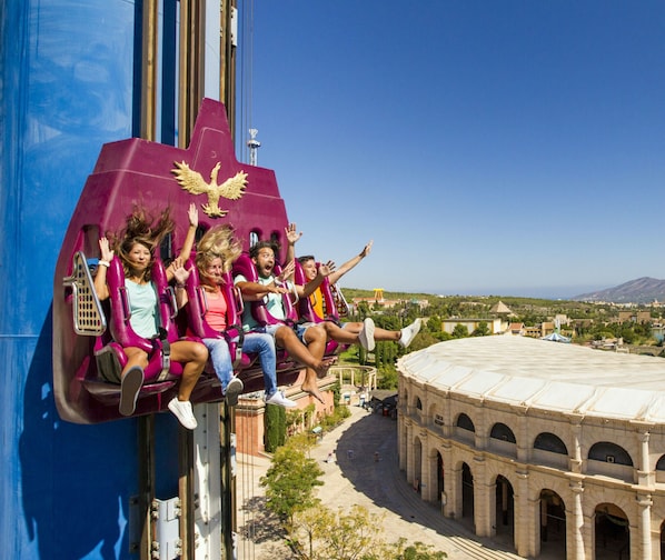 A roller coaster with passengers in mid-air, overlooking a historic building with arches.
