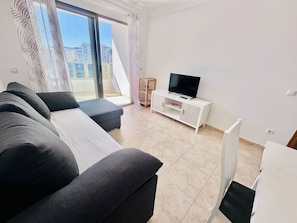 A modern living room with a sofa, a TV on a white cabinet, and a balcony with a view of buildings.
