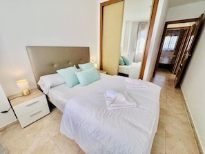 A neatly made bed with white linens and light blue pillows, a bedside lamp, and a wooden nightstand with drawers.