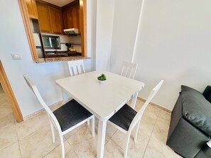 A small dining area with a white table and four chairs, a small potted plant on the table, and a kitchen in the background. - A1089 - Sunny apartment with pool view (Villajoyosa)