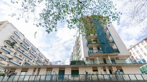 Un immeuble à plusieurs étages doté de balcons, une devanture de magasin avec des enseignes indiquant « Beria » et « Da Carlo », et un pont piétonnier avec des garde-corps verts.