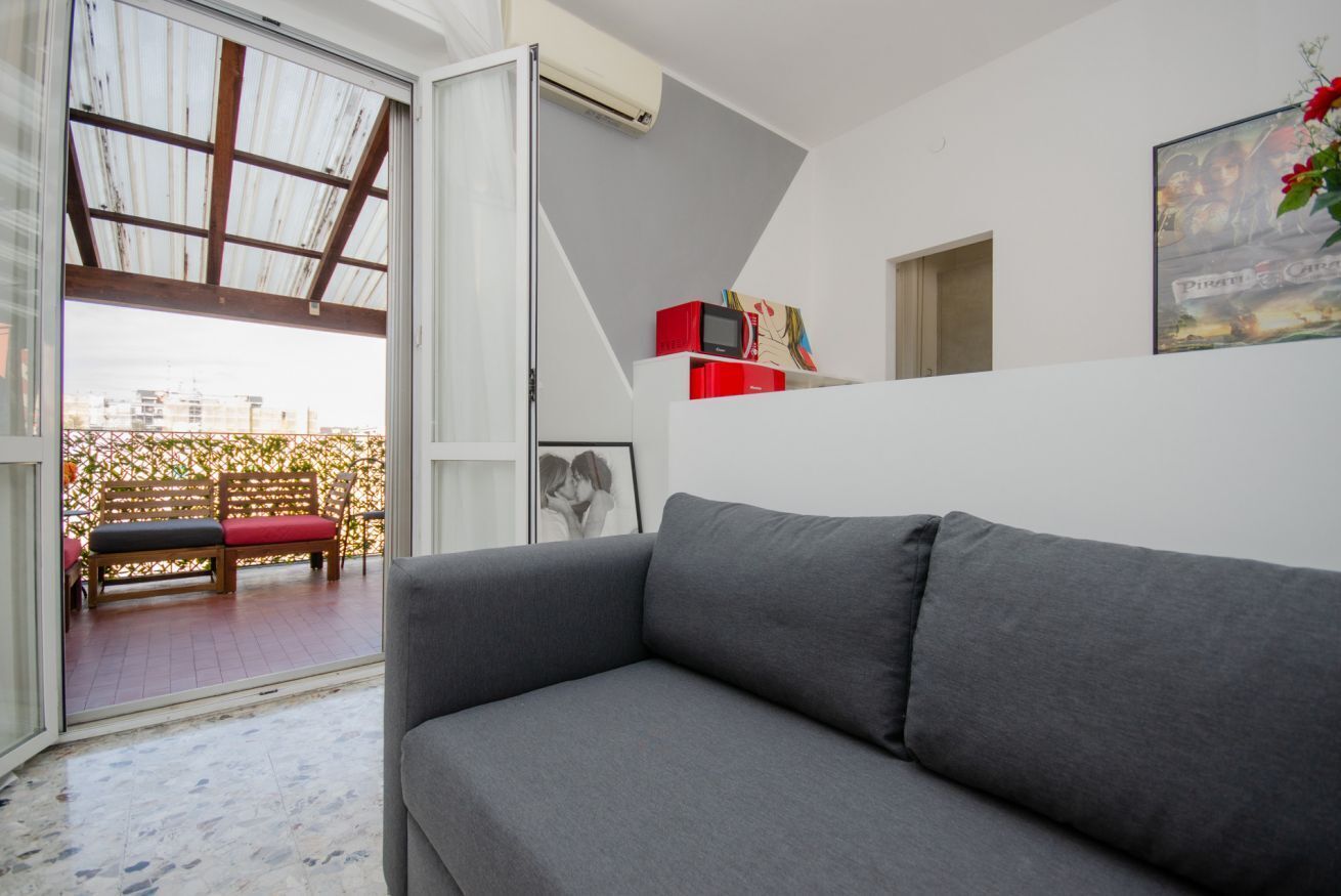 Uma sala de estar moderna com um sofá cinza, um suporte de TV vermelho e uma janela grande com vista para uma varanda com um banco vermelho.