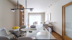 A modern living room with a glass dining table, grey chairs, and a sofa. There is a bookshelf and a window with curtains. - Maximus Design By House and People (Braga)