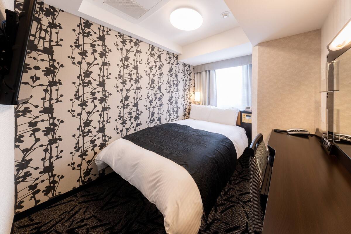 Une chambre d’hôtel avec un lit, un bureau, un miroir, une télévision et une fenêtre avec des rideaux.