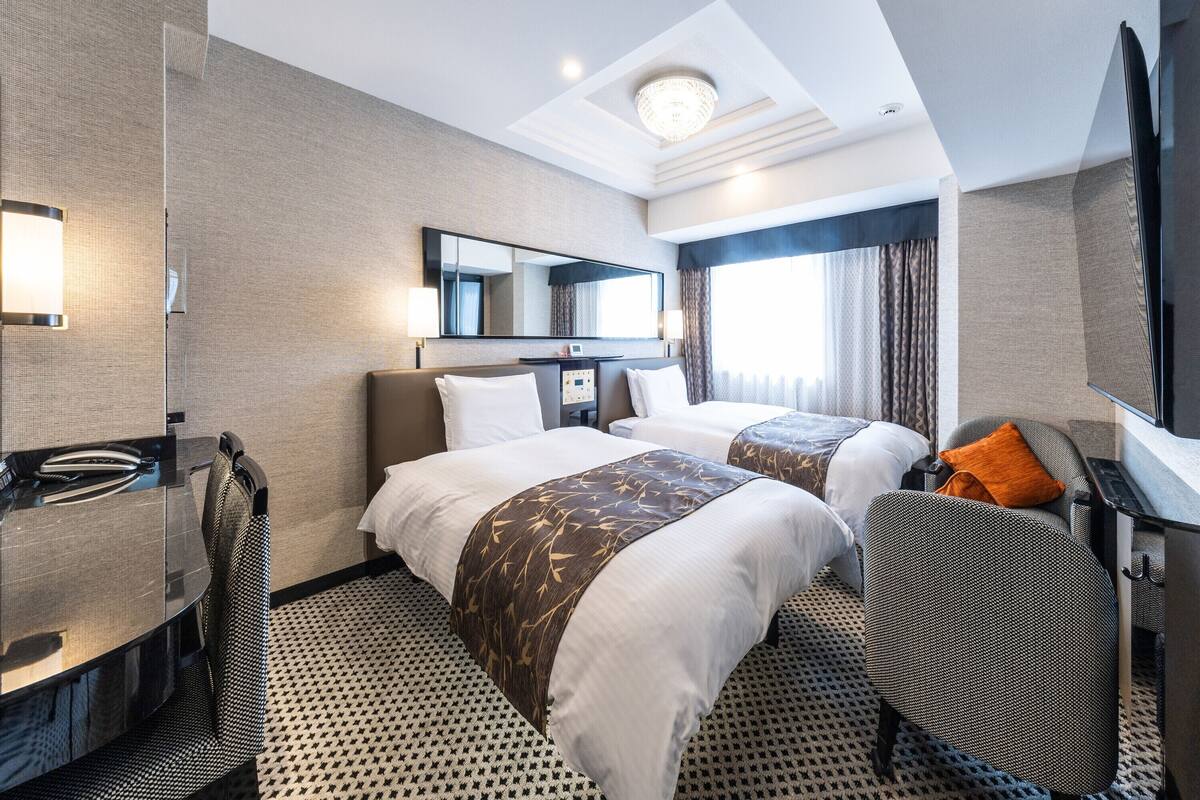 Une chambre d’hôtel avec deux lits, un bureau, une chaise, un miroir et une grande fenêtre avec des rideaux.