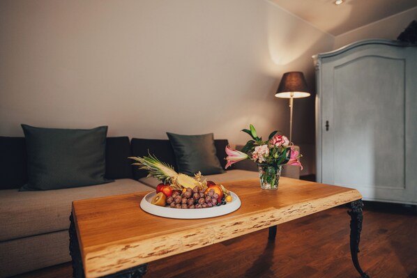 A wooden table with a fruit platter and a vase of flowers in a living room with a sofa and a lamp.