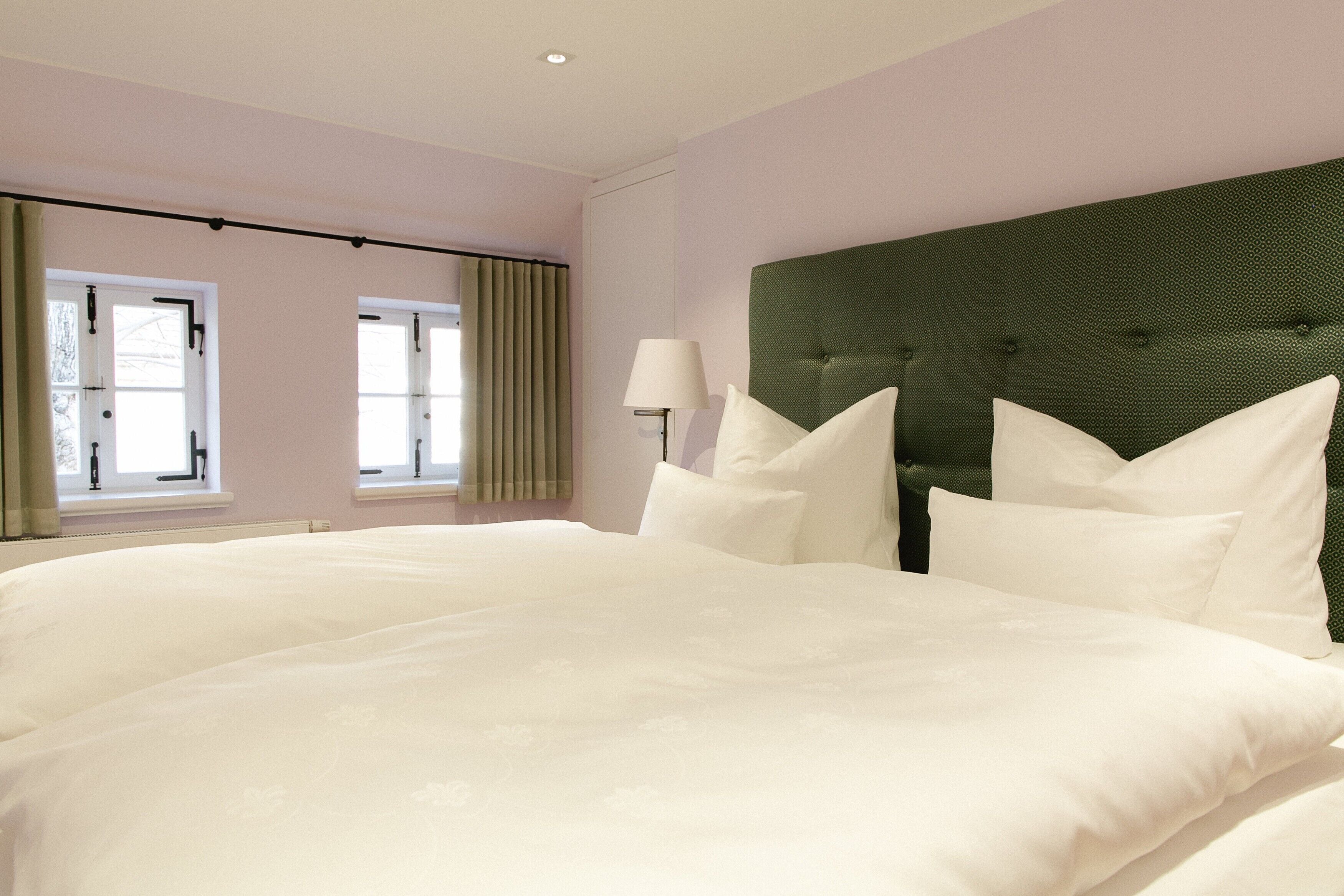 Una habitación de hotel con una cama grande, un cabecero tapizado en verde y dos ventanas con cortinas.