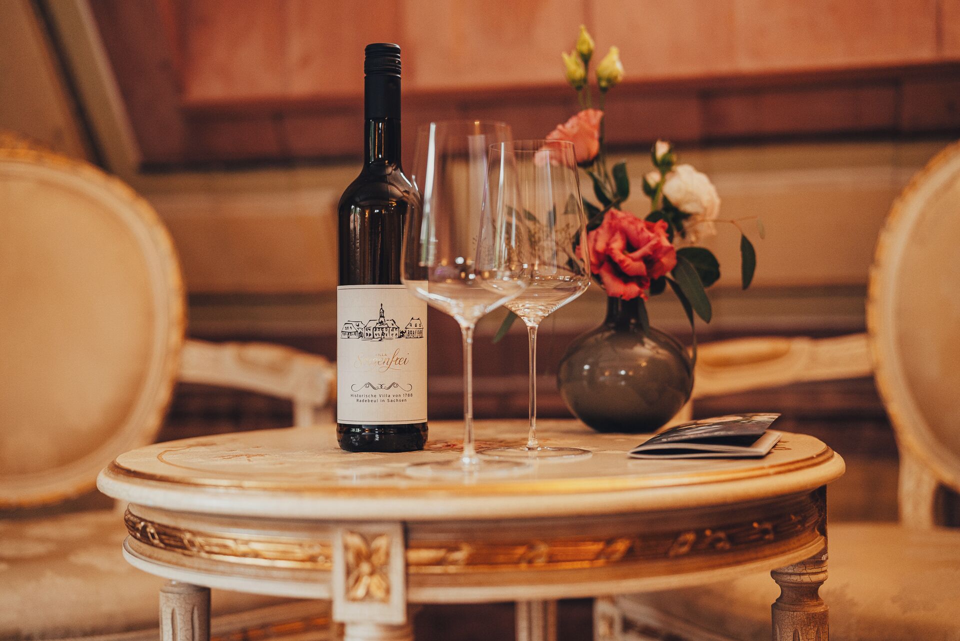 A bottle of wine, two glasses, and a vase with flowers on a round table.