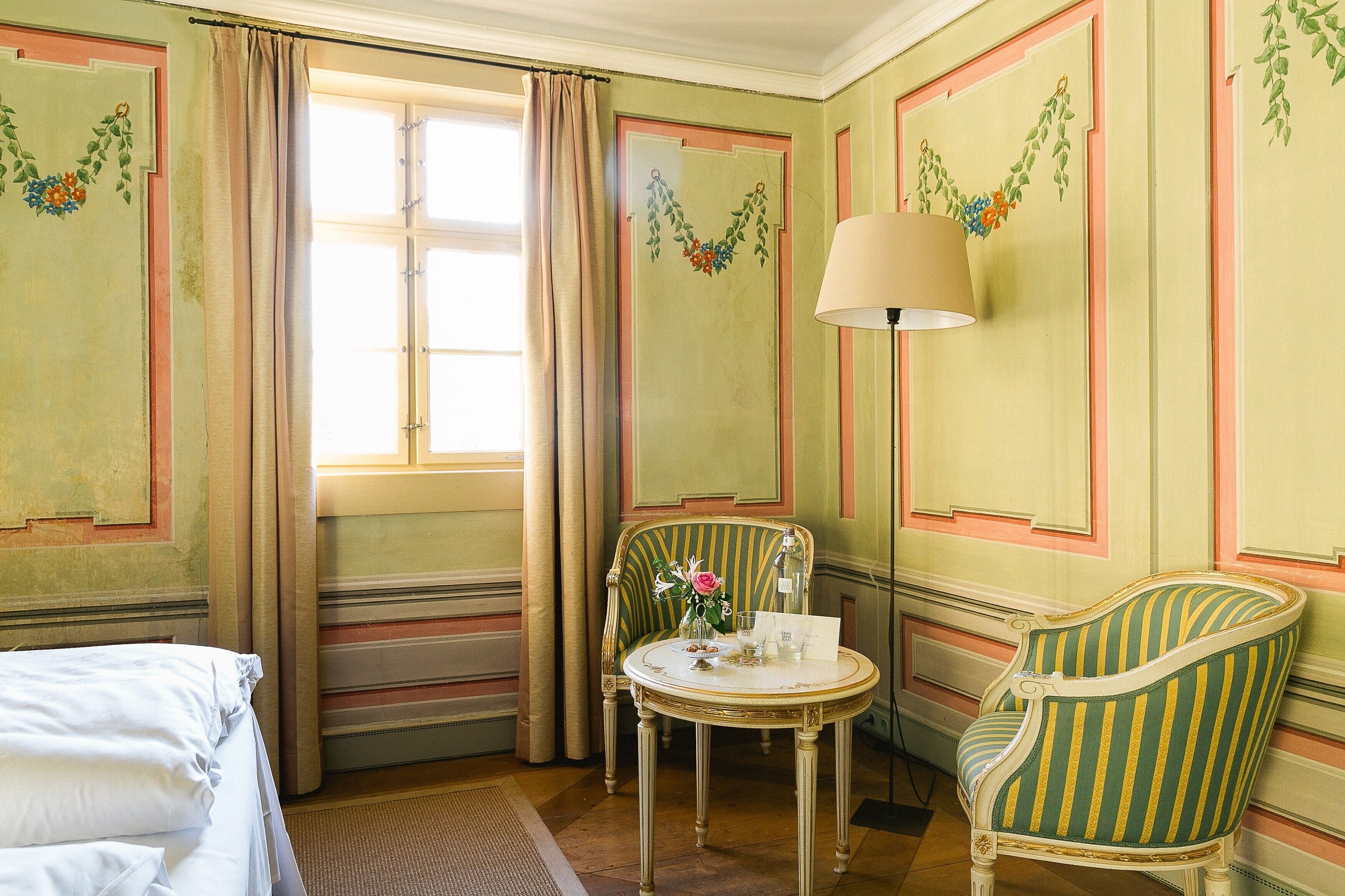 A room with a bed, a chair, a table with a vase of flowers, and a lamp.