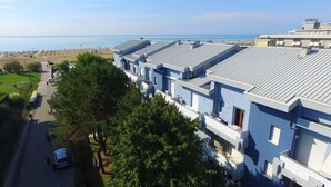 A row of modern, light blue buildings with flat roofs, likely part of a hotel or resort, adjacent to a beach with a clear view of the sea. - Charming studio apartment by the sea (Bibione)