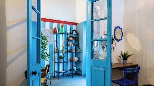 A room with a blue door, a wooden desk, a blue chair, and a metal shelf unit with various items.