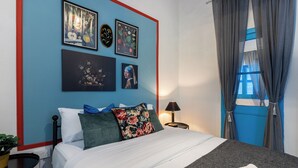 A bedroom with a bed, pillows, a nightstand, a lamp, and framed botanical prints on the wall.