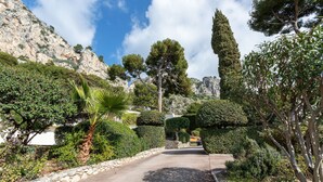 A paved pathway flanked by neatly trimmed hedges and trees, leading towards a rocky mountain backdrop. - LA SOLEILLADE VI4451 By Riviera Holiday Homes (Eze)