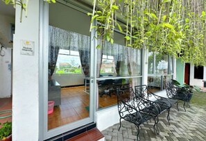 An outdoor seating area with metal chairs and tables, a glass door entrance, and hanging plants. - M&S Stay Syariah (Sleman)
