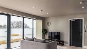 Een moderne woonkamer met een grijze bank, een flatscreen televisie en een grote glazen schuifdeur die leidt naar een buitenruimte.