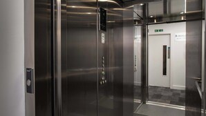 Een lift met een digitaal scherm, een nooduitgangsbord en een deur met een naambordje.