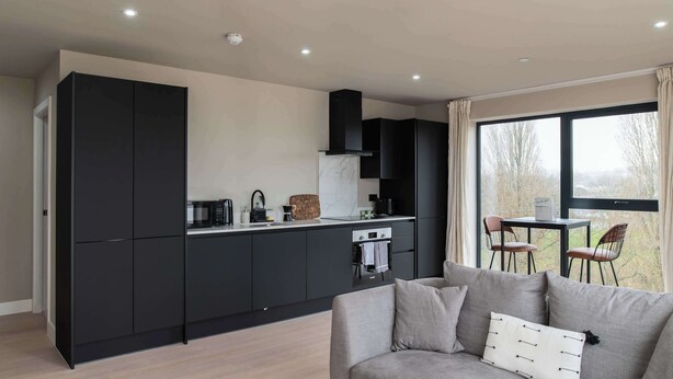 Een moderne keuken met zwarte kastjes, een grijze bank en een raam met uitzicht op bomen.