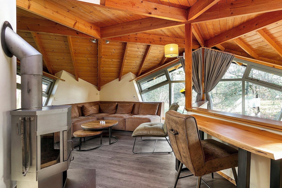 Interior acogedor de una cabaña con techo de madera, chimenea de piedra, sofá, mesa de comedor y ventanas amplias con vistas al exterior.