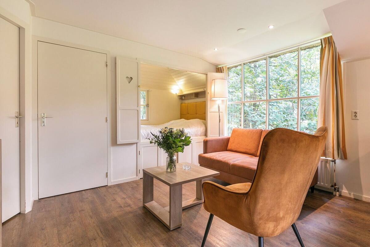 Una habitación moderna con piso de madera, un sillón de cuero marrón, una mesita con un florero, una ventana con cortinas y una cama al fondo.