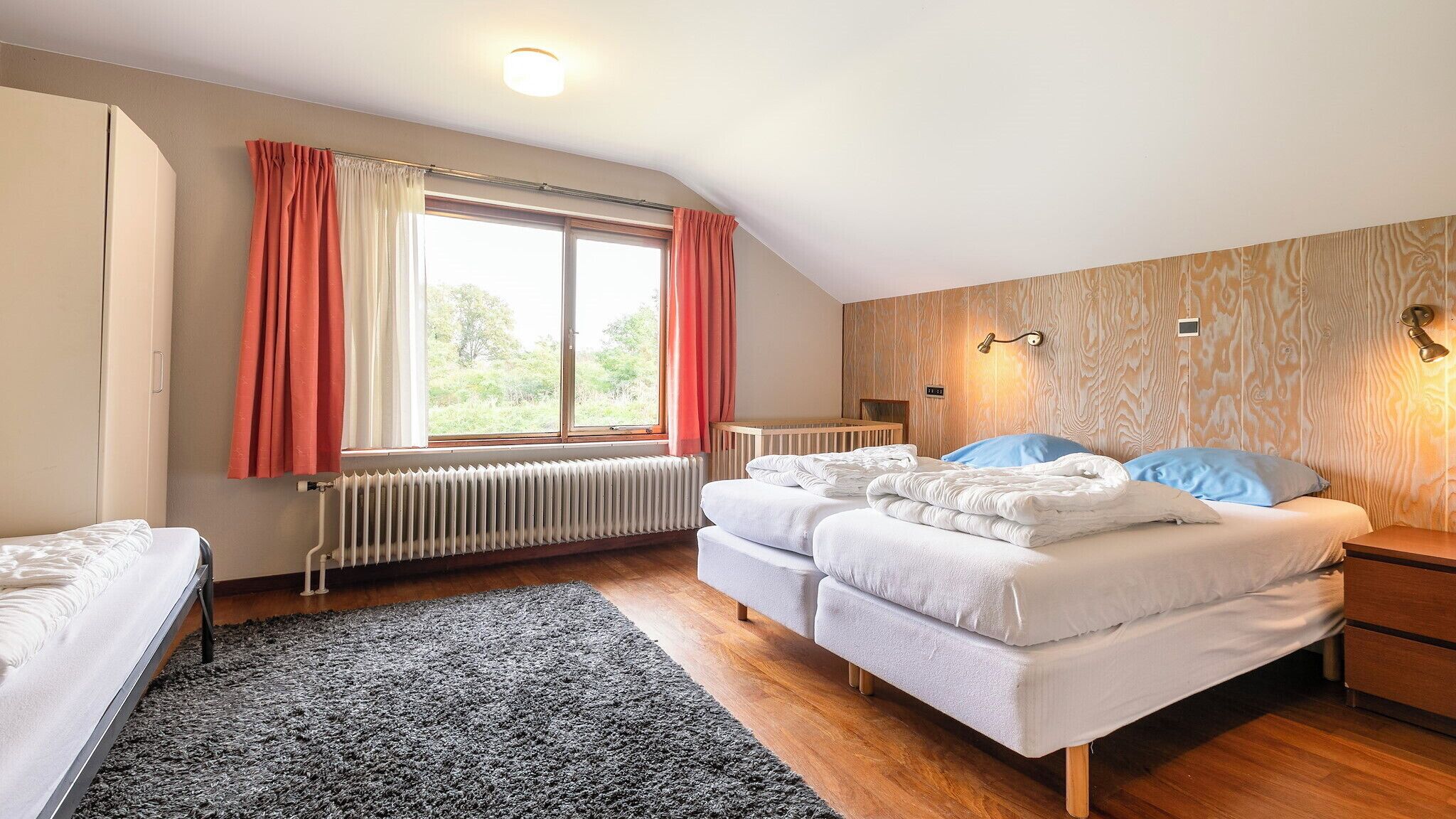 A bedroom with two beds, a window with curtains, a radiator, a wooden headboard, and a nightstand.