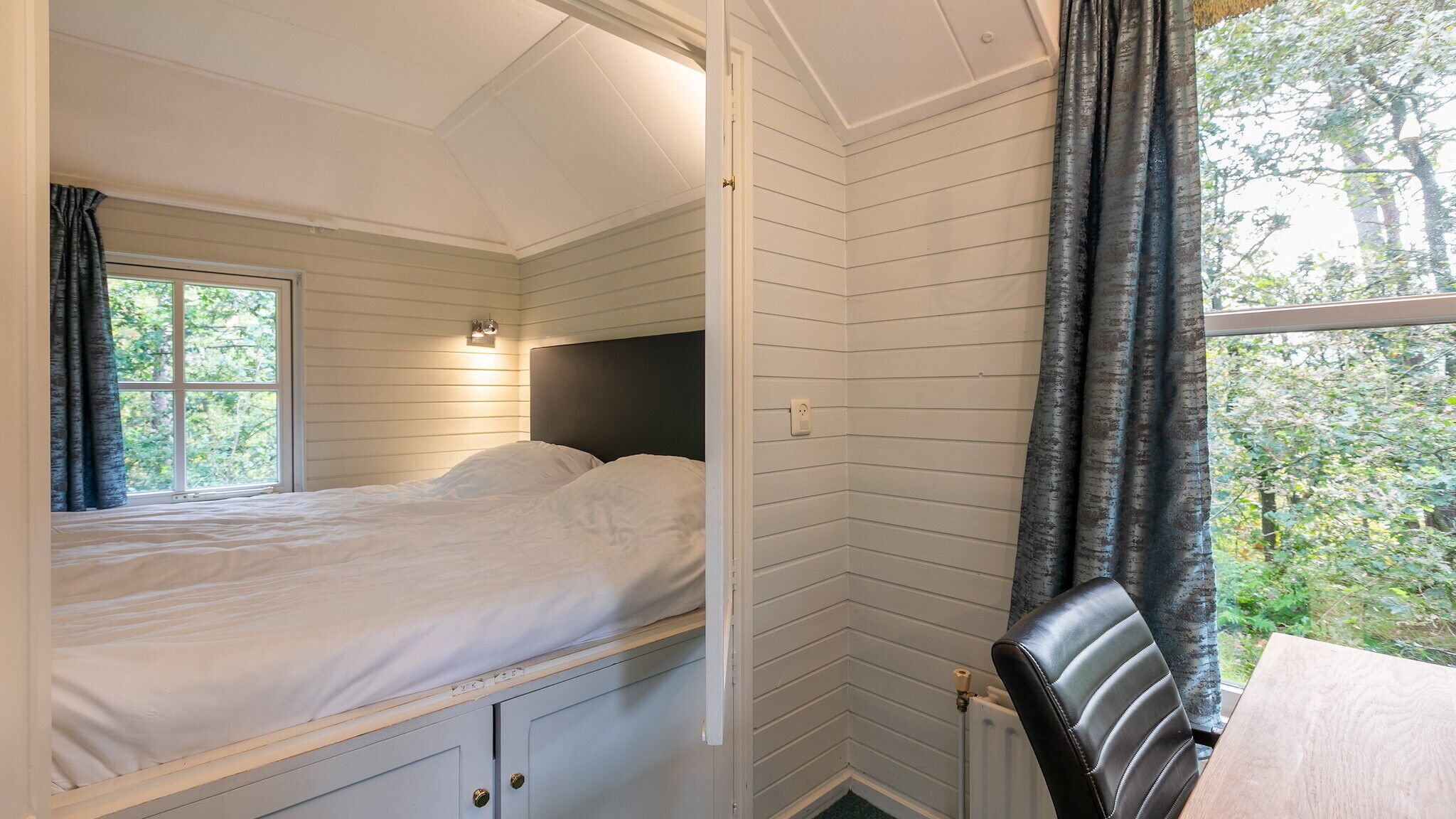 A bedroom with a bed, a nightstand, a window with curtains, and a view of greenery outside.