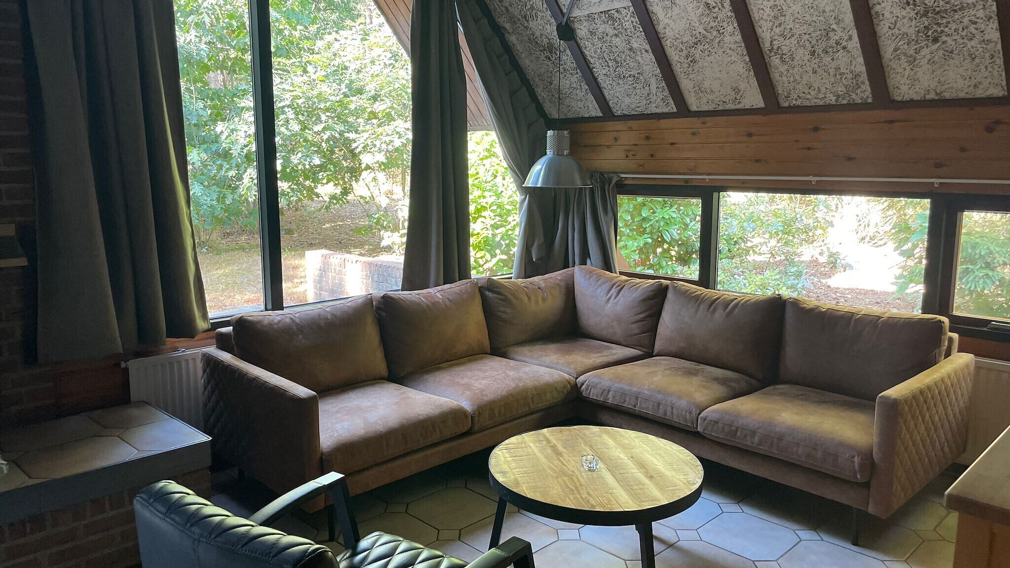 A cozy living room with a sectional sofa, a round coffee table, and large windows offering a view of greenery outside.