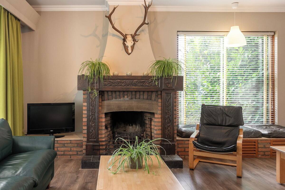 Una sala de estar con chimenea, cortinas verdes, televisor, sofá, sillón y ventana con persianas.