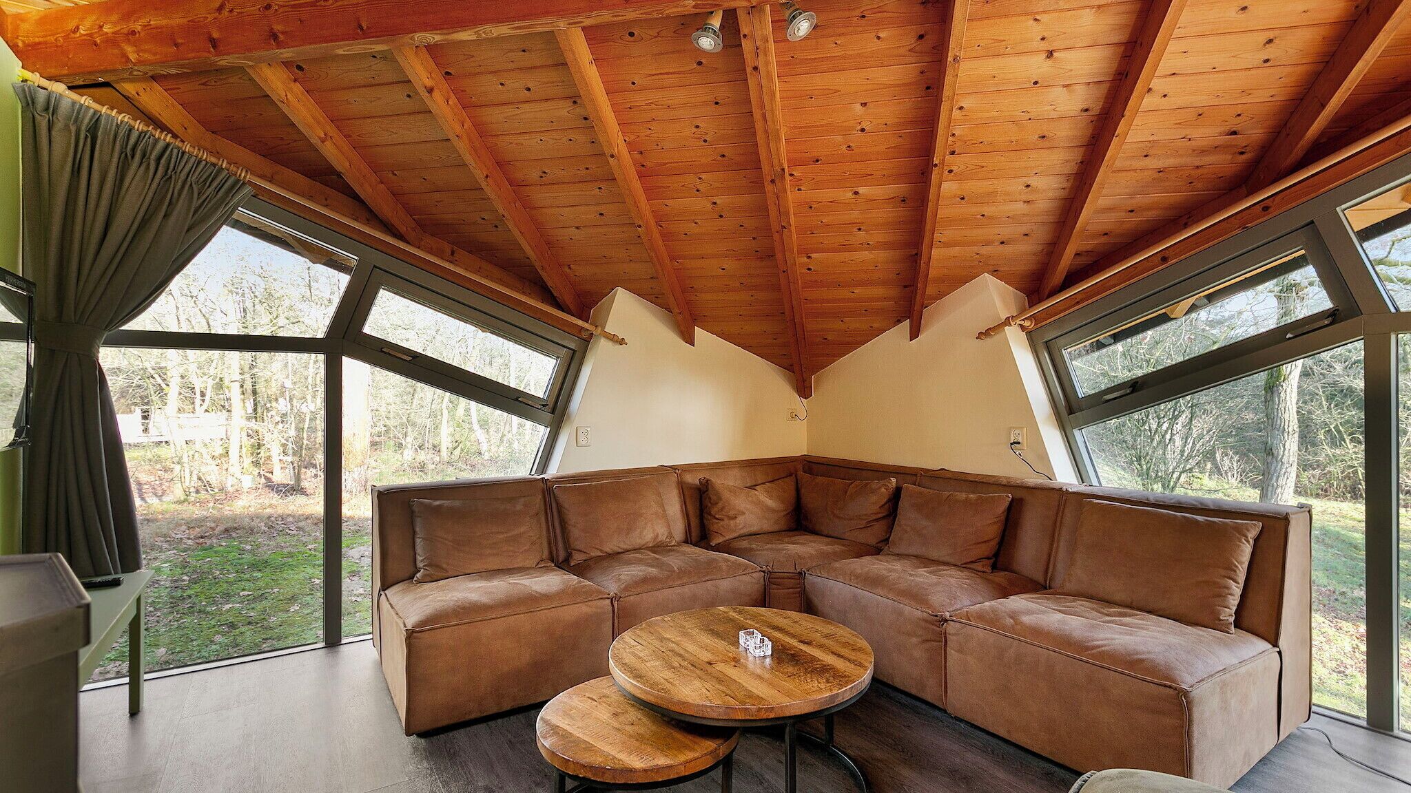 A cozy corner with a brown leather sectional sofa, two wooden round tables, and large windows offering a view of a forested area.