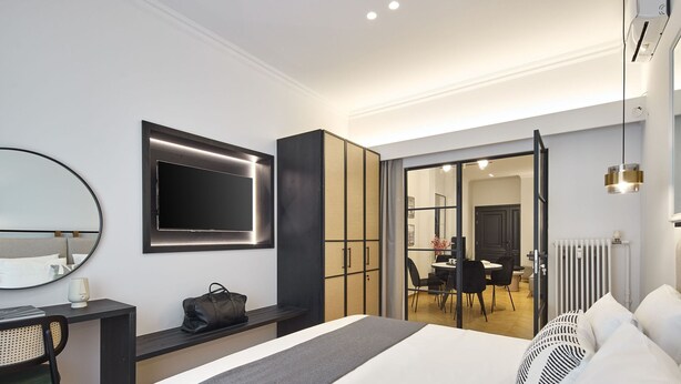 A modern hotel room with a large bed, a desk with a chair, a TV mounted on the wall, and a sliding door leading to a dining area. - The Athenian Chic & Elegant Apartments in Kolonaki (Athens)
