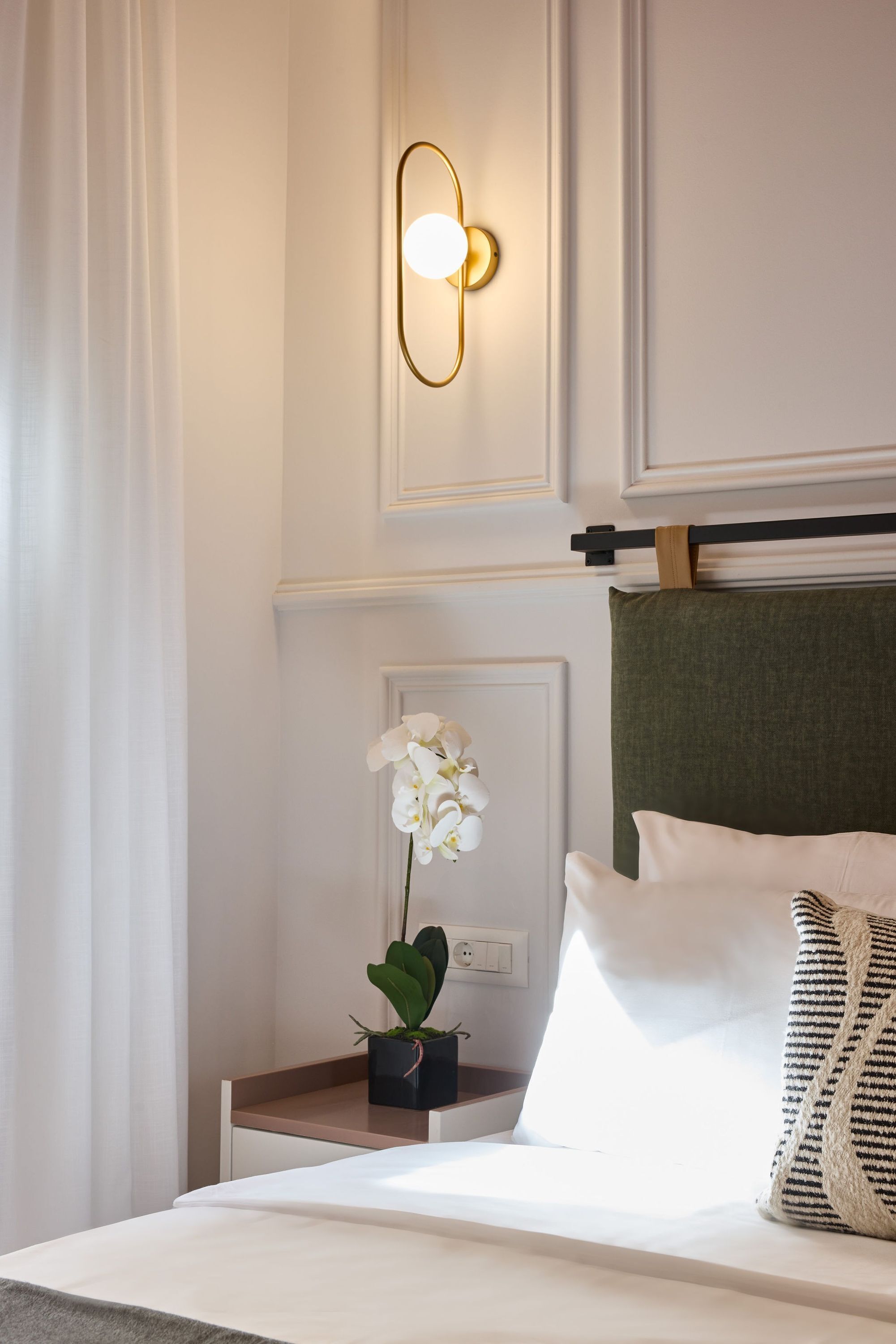 A hotel room with a neatly made bed, a bedside table with a potted orchid, a wall-mounted light fixture, and a green upholstered headboard.
