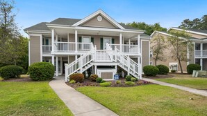 A two-story residential building with a front porch, a staircase leading to the second floor, and a well-maintained lawn. - Heart of Pawleys Island 3BR Golf Condo Across from Pool (Pawleys Island)
