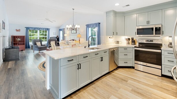 A modern kitchen with grey cabinets, a large island, stainless steel appliances, and a dining area with a grey sofa and blue curtains. - Heart of Pawleys Island 3BR Golf Condo Across from Pool (Pawleys Island)