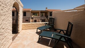 Un patio al aire libre con dos sillas metálicas verdes y una mesa, suelo de baldosas y un edificio con techo de tejas al fondo.
