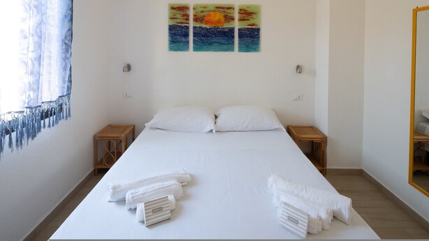 A neatly made bed with white linens and towels, a wall-mounted painting of a sunset over the ocean, and a wooden nightstand on each side of the bed. - Sabbia Marina (Budoni)