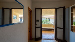 A hallway with a wooden floor leading to an open door revealing a balcony with a chair and a view of a building with a tiled roof. - Resinosa in Agrustos (Budoni)