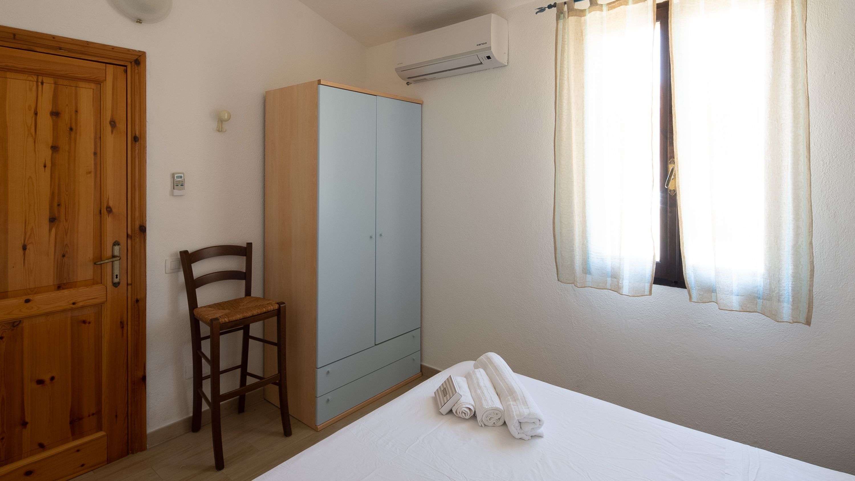 A bedroom with a bed, a wooden chair, a wardrobe, and a window with curtains.