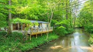A cabin with a deck overlooking a river, surrounded by dense greenery. - Splash at the Creek, roast marshmallows & play ping-pong together – Wade 'N Bear (Sevierville)