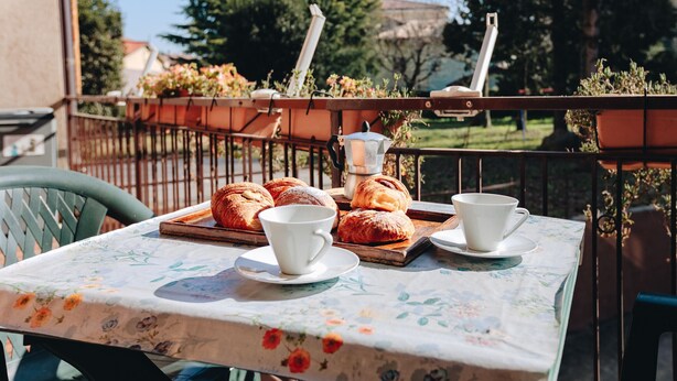 Una mesa con mantel floral, dos tazas blancas y una bandeja de pasteles en un balcón.