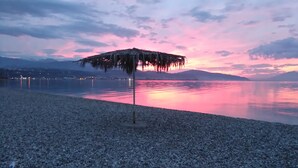Een kiezelstrand met een rieten parasol, kalm water en verafgelegen bergen.