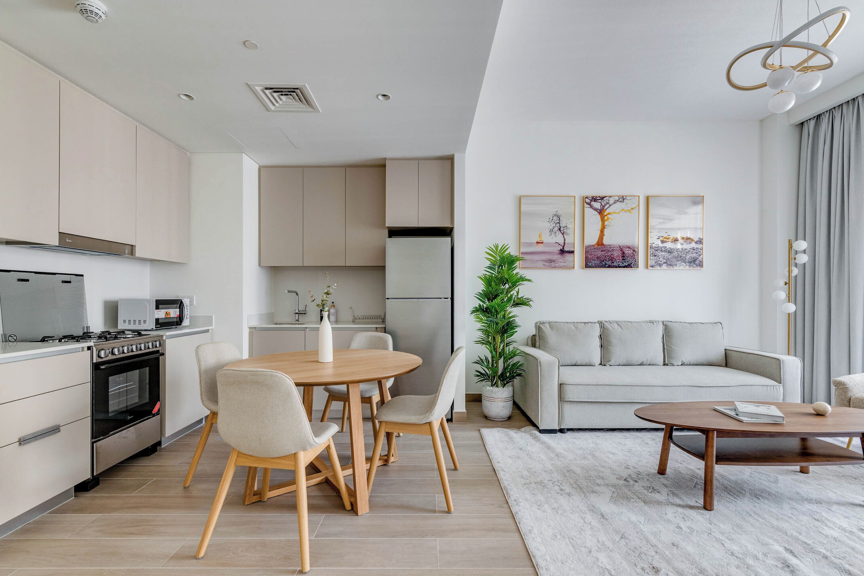 A modern kitchen with built-in appliances, a dining area with a round table and chairs, a living room with a sofa and a coffee table, and a large window with curtains.