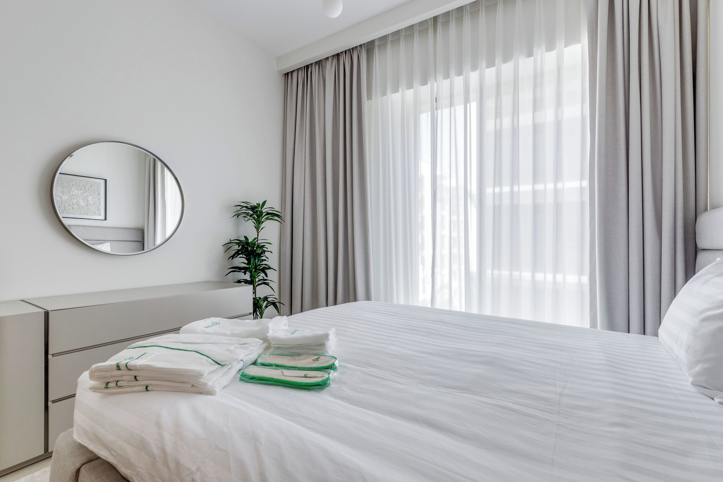 A neatly made bed with white linens and green towels, a bedside plant, a round mirror, and a window with sheer curtains.