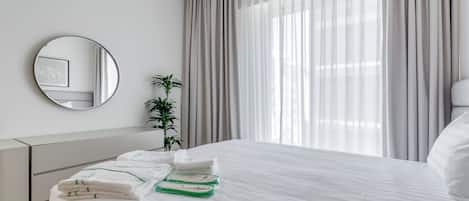 A neatly made bed with white linens and green towels, a bedside plant, a round mirror, and a window with sheer curtains.