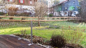 Eine Holzterrasse mit Metallgeländer, umgeben von einer Rasenfläche mit vereinzelter Bepflanzung und Wohnhäusern im Hintergrund.