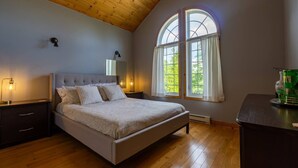 A bedroom with a bed, bedside tables, a nightstand, a window with curtains, and a wooden ceiling.