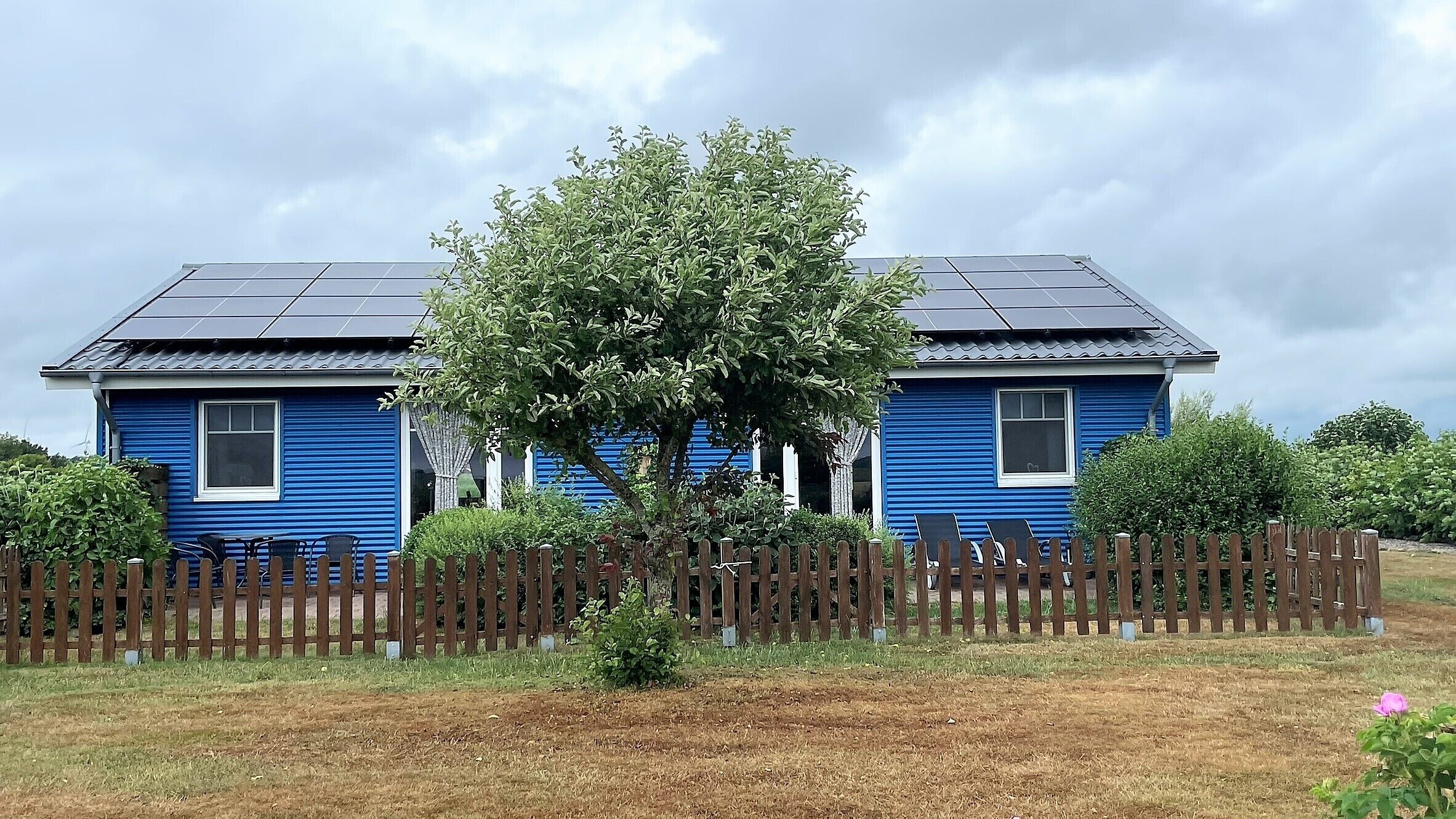 Ein blaues Haus mit Solaranlage, Holzzaun und Baum davor.