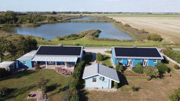 Zona residencial con dos casas azules que tienen paneles solares, un estanque y aerogeneradores al fondo.