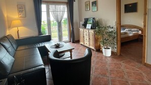 Sala de estar con sofá, una mesa de centro y televisor sobre un mueble de madera. En la habitación contigua hay una cama y una planta en maceta en la esquina.