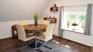 A dining area with a round wooden table, four chairs, a vase with green plants, and a window with curtains. - Experience Tranquility at Ferienwohnung Vadders Hof in Risum-Lindholm! (Risum-Lindholm)