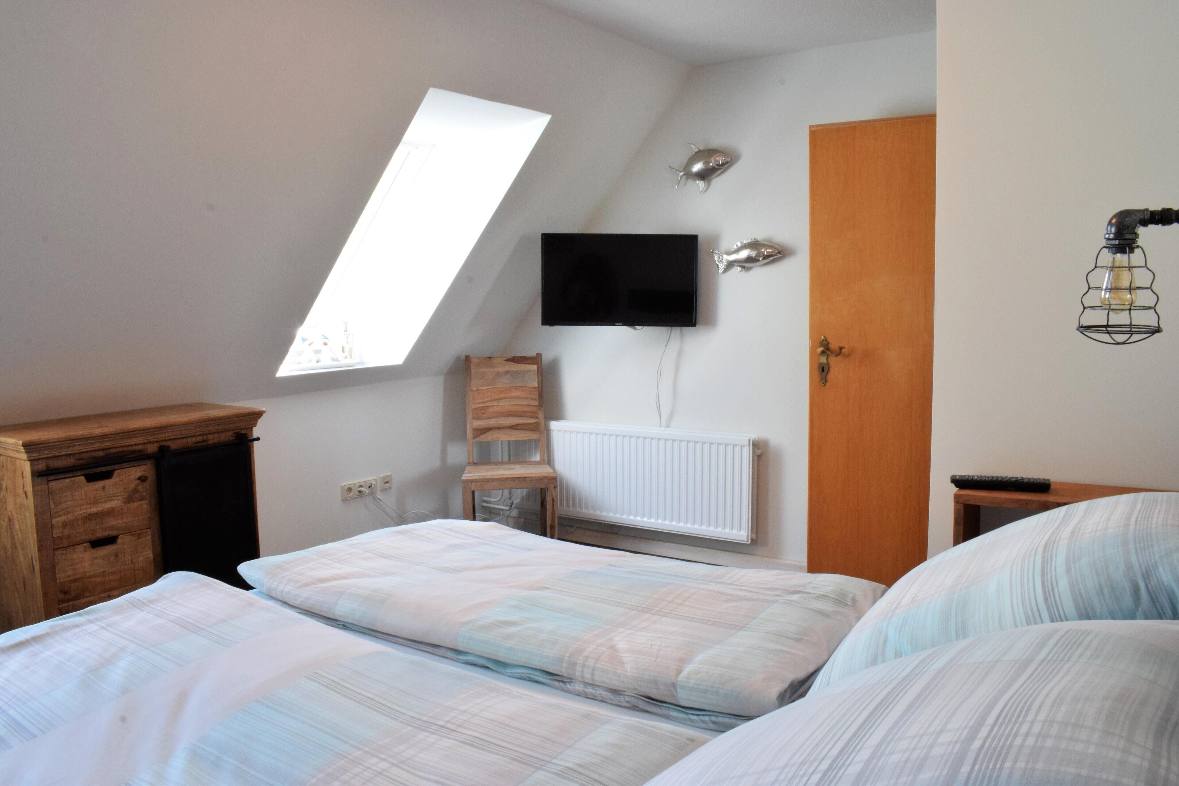 A bedroom with a sloped ceiling, a TV mounted on the wall, a wooden dresser, a chair, and a bed with a striped blanket.